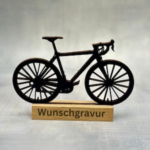 May include: A black bicycle silhouette, featuring detailed spokes and handlebars, is mounted on a wooden base. The base has the word "Wunschgravur" engraved on it. The bicycle is a decorative item.