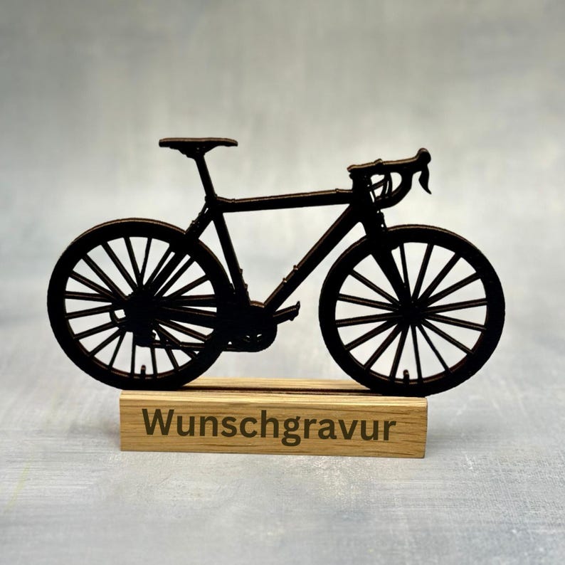 May include: A black bicycle silhouette, featuring detailed spokes and handlebars, is mounted on a wooden base. The base has the word "Wunschgravur" engraved on it. The bicycle is a decorative item.