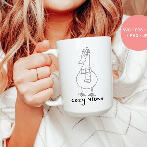 K&ouml;nnte beinhalten: Wei&szlig;e Keramiktasse mit einer schwarzen Strichzeichnung einer Gans, die einen Hut und einen Schal tr&auml;gt. Der Text "cozy vibes" ist unter der Gans aufgedruckt.