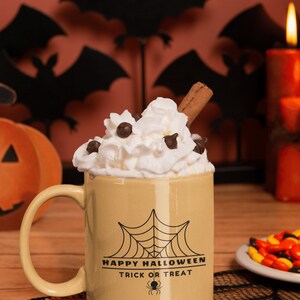 K&ouml;nnte beinhalten: Eine beige Keramiktasse mit einem schwarzen Spinnennetz und dem Text "Happy Halloween Trick or Treat" auf der Seite. Die Tasse ist gef&uuml;llt mit einem hei&szlig;en Getr&auml;nk, das mit Schlagsahne, Schokoladenst&uuml;ckchen und einem Zimtstange garniert ist.