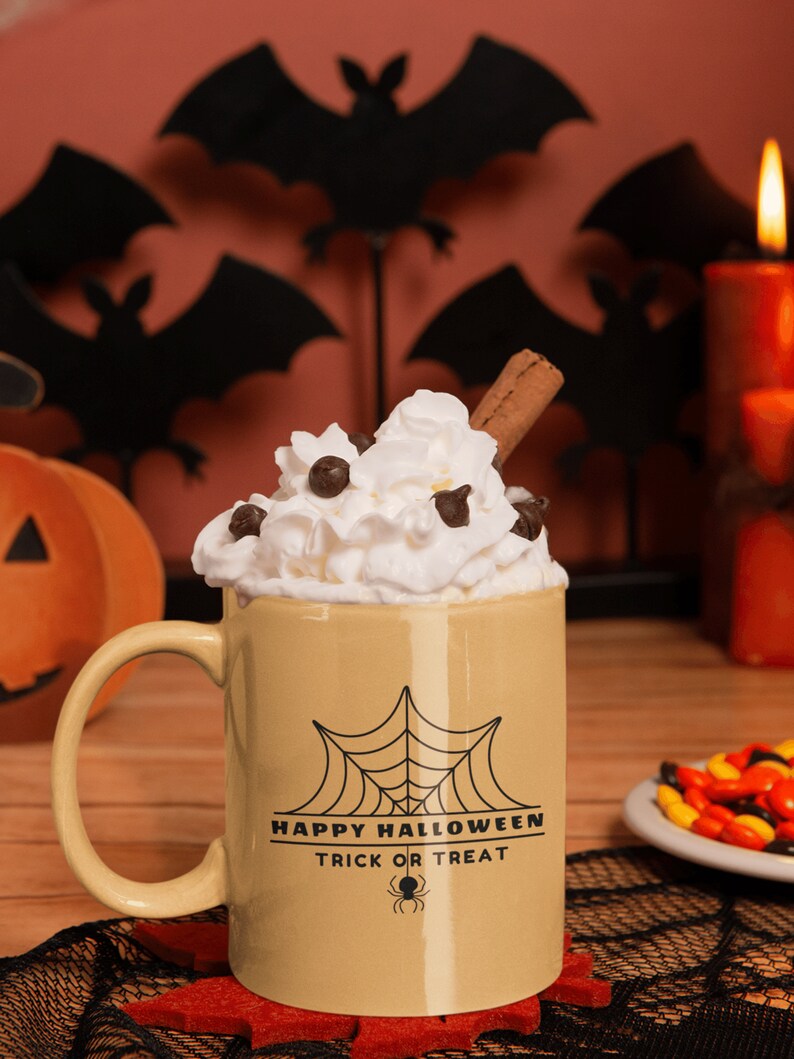 K&ouml;nnte beinhalten: Eine beige Keramiktasse mit einem schwarzen Spinnennetz und dem Text "Happy Halloween Trick or Treat" auf der Seite. Die Tasse ist gef&uuml;llt mit einem hei&szlig;en Getr&auml;nk, das mit Schlagsahne, Schokoladenst&uuml;ckchen und einem Zimtstange garniert ist.