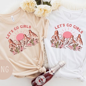 May include: Two white t-shirts with a desert landscape graphic and the text "Let's Go Girls". The t-shirts are laid out on a white surface with a bottle of sparkling wine.