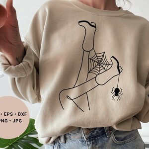 May include: A beige sweatshirt with a black line drawing of a person's legs tangled in a spider web with a spider hanging from the web.