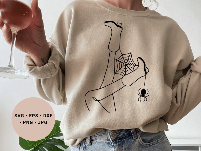 May include: A beige sweatshirt with a black line drawing of a person's legs tangled in a spider web with a spider hanging from the web.