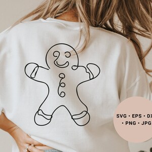 Könnte beinhalten: Ein weißes Sweatshirt mit einer schwarzen Linienzeichnung eines Lebkuchenmanns auf der Vorderseite. Die Zeichnung ist einfach und zeigt ein lächelndes Gesicht, Knöpfe und ausgestreckte Arme. Der Text "SVG - EPS - DXF - PNG - JPG" befindet sich auf einem rosa Kreis in der unteren rechten Ecke des Bildes.