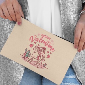 May include: A beige canvas zippered pouch with a pink and white illustration of a cowboy riding a horse with the text "Howdy Valentine" and pink hearts.