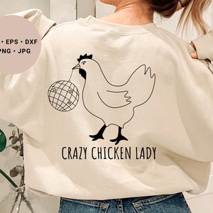 Könnte beinhalten: Ein beiger Pullover mit einer schwarzen Strichzeichnung eines Huhns, das eine Discokugel hält. Der Text "Crazy Chicken Lady" ist unter dem Huhn gedruckt.
