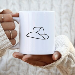 May include: White ceramic mug with a black line drawing of a cowboy hat.
