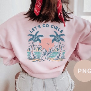 K&ouml;nnte beinhalten: Hellrosa Sweatshirt mit dem Text "LET'S GO GIRLS" &uuml;ber einer Strandszene mit Palmen, Sonnenuntergang und den Beinen einer Person in Cowboystiefeln. Das Sweatshirt hat eine hellrosa Farbe.