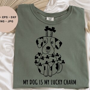 Könnte beinhalten: Ein grünes T-Shirt mit einer schwarz-weißen Illustration eines Hundes, der ein Kleeblatt-Stirnband und eine Kleeblatt-Girlande trägt. Der Text "MY DOG IS MY LUCKY CHARM" ist unter der Illustration aufgedruckt.