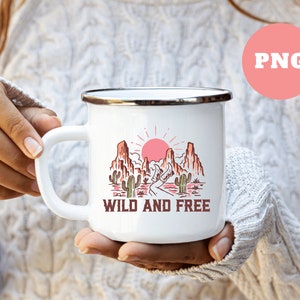 Puede incluir: Taza de esmalte blanco con un gr&aacute;fico de un paisaje des&eacute;rtico con un sol rojo, monta&ntilde;as y cactus. El texto "WILD AND FREE" est&aacute; impreso debajo del gr&aacute;fico.