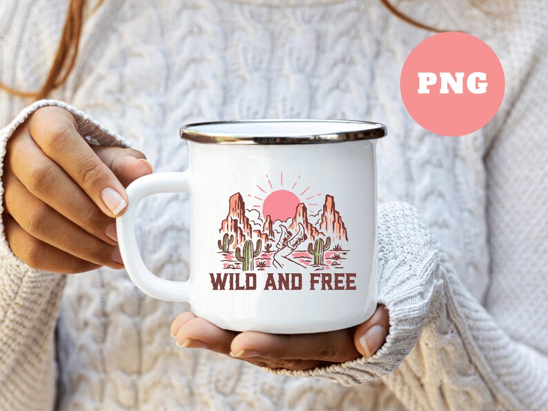 Puede incluir: Taza de esmalte blanco con un gr&aacute;fico de un paisaje des&eacute;rtico con un sol rojo, monta&ntilde;as y cactus. El texto "WILD AND FREE" est&aacute; impreso debajo del gr&aacute;fico.