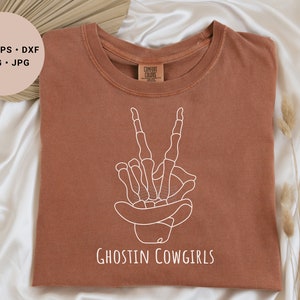 May include: A brown t-shirt with a white outline of a skeleton hand making a peace sign wearing a cowboy hat. The text "Ghostin Cowgirls" is printed below the hand.