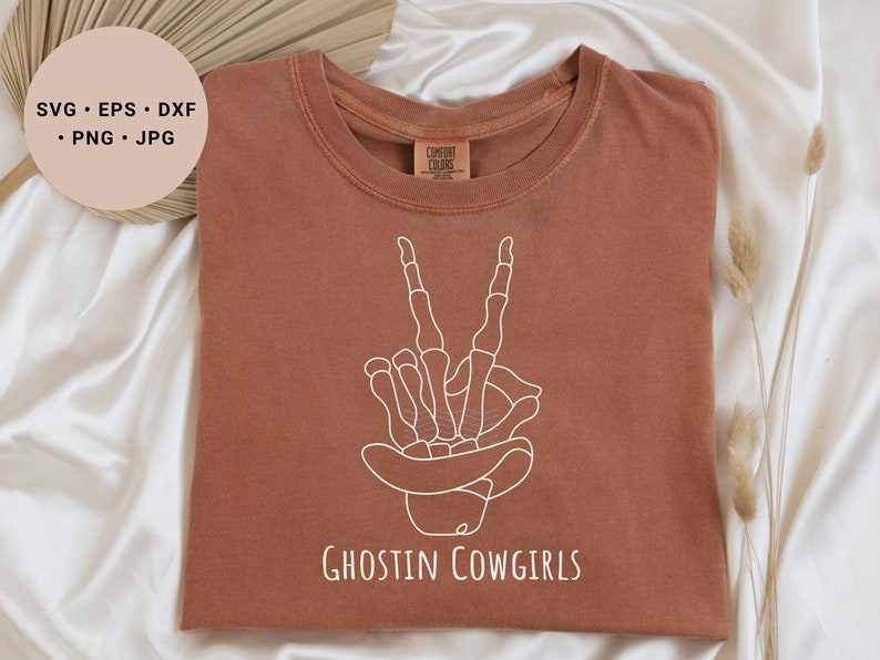 May include: A brown t-shirt with a white outline of a skeleton hand making a peace sign wearing a cowboy hat. The text "Ghostin Cowgirls" is printed below the hand.