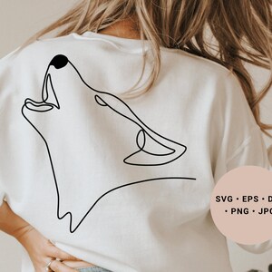 Puede incluir: Una sudadera blanca con un dibujo de línea negra de un lobo aullando en la espalda. El texto "SVG - EPS - DXF - PNG - JPG" está impreso en la parte inferior derecha de la sudadera.