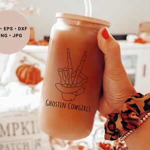 May include: A brown glass tumbler with a straw, featuring a black line drawing of a skeleton hand making a peace sign wearing a cowboy hat. The tumbler has the text "Ghostin Cowgirls" printed on it.