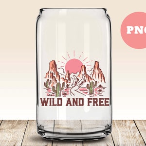 Puede incluir: Una lata de vidrio transparente con un dise&ntilde;o de paisaje des&eacute;rtico con un sol rojo, monta&ntilde;as, cactus y un camino con el texto "WILD AND FREE".