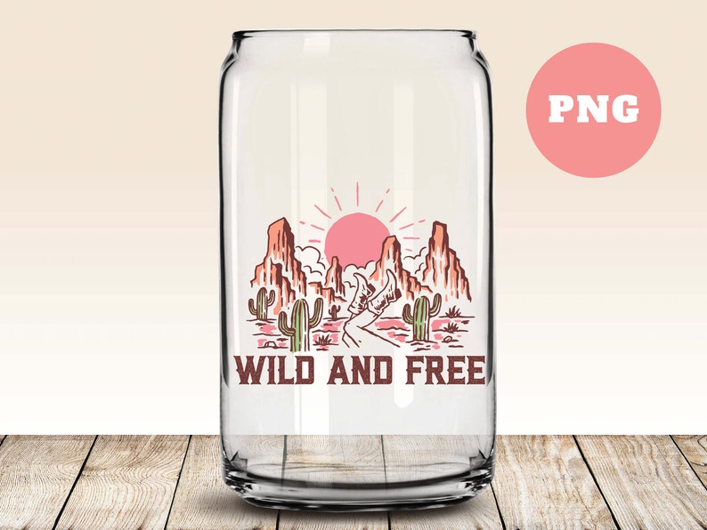 Puede incluir: Una lata de vidrio transparente con un dise&ntilde;o de paisaje des&eacute;rtico con un sol rojo, monta&ntilde;as, cactus y un camino con el texto "WILD AND FREE".