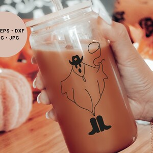 May include: A clear glass tumbler with a brown beverage inside. A black and white line drawing of a ghost wearing a cowboy hat and boots is on the side of the glass. The glass is being held by a hand wearing a bracelet with a small white ghost charm.