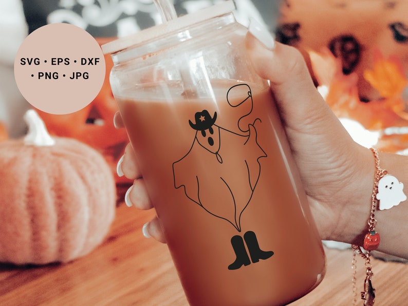 May include: A clear glass tumbler with a brown beverage inside. A black and white line drawing of a ghost wearing a cowboy hat and boots is on the side of the glass. The glass is being held by a hand wearing a bracelet with a small white ghost charm.
