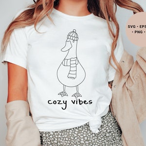 K&ouml;nnte beinhalten: Wei&szlig;es T-Shirt mit einer schwarzen Strichzeichnung eines Entchens mit Hut und Schal. Der Text "cozy vibes" ist unter dem Entchen gedruckt.