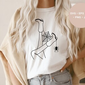 May include: A white t-shirt with a black line drawing of a person's legs tangled in a spider web with a spider hanging from the web.