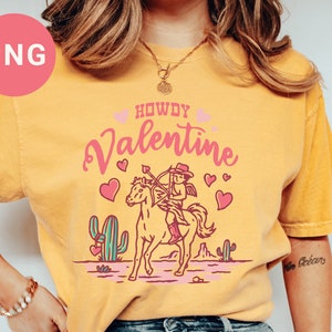 May include: A yellow t-shirt with the text "Howdy Valentine" in pink lettering. A cartoon cupid riding a horse with an arrow is in the center of the shirt. The cupid is wearing a cowboy hat and boots.