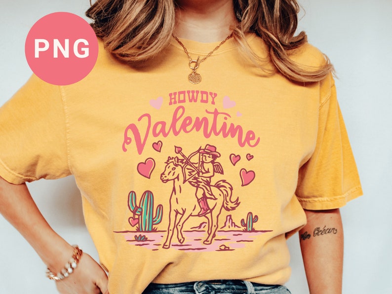 May include: A yellow t-shirt with the text "Howdy Valentine" in pink lettering. A cartoon cupid riding a horse with an arrow is in the center of the shirt. The cupid is wearing a cowboy hat and boots.