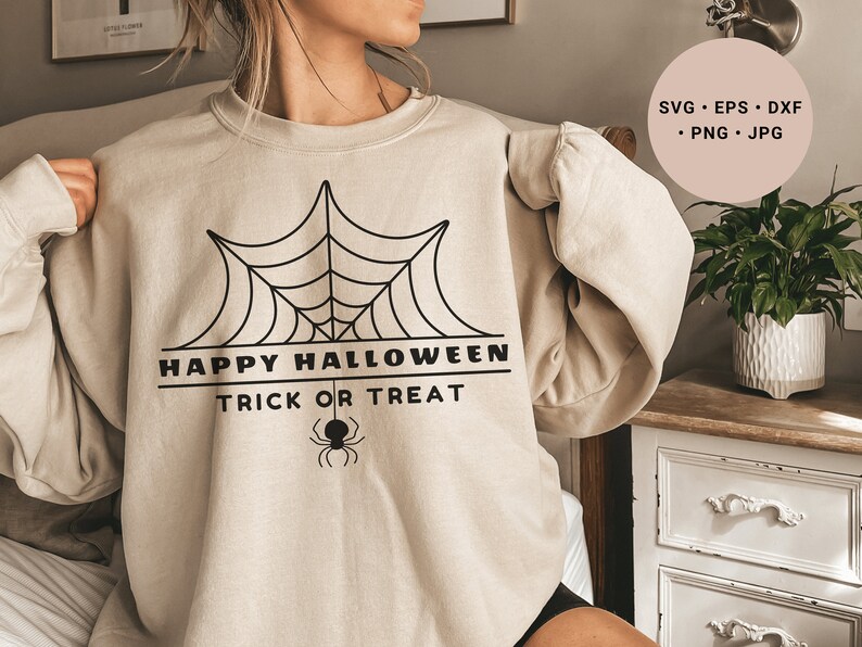 K&ouml;nnte beinhalten: Ein beiger Pullover mit einem schwarzen Spinnennetz-Design und dem Text "Happy Halloween Trick or Treat".