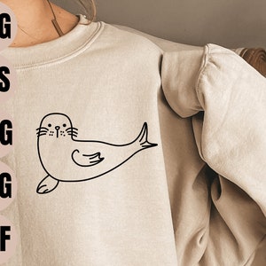 May include: A black and white line drawing of a seal on a beige sweatshirt. The seal is facing forward with its flippers outstretched. The drawing is simple and stylized.