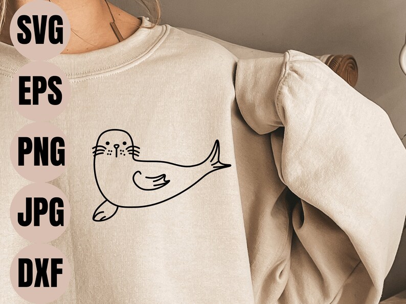 May include: A black and white line drawing of a seal on a beige sweatshirt. The seal is facing forward with its flippers outstretched. The drawing is simple and stylized.