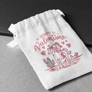 May include: White drawstring bag with a pink and white illustration of a cowgirl riding a horse with the text "Howdy Valentine" and pink hearts.