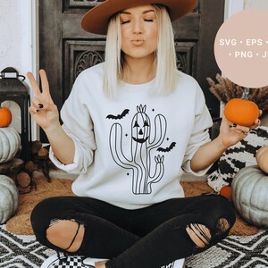 May include: A white sweatshirt featuring a black line drawing of a cactus with a jack-o'-lantern face and bats flying around it. The text "SVG - EPS - DXF - PNG - JPG" is in a peach-coloured circle in the top right corner of the image.