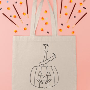 May include: A white canvas tote bag with black text that says "Howdy Pumpkin" and a black line drawing of a person's legs sticking out of a jack-o'-lantern. The bag is surrounded by candy corn, bats, and spiders on a pink background.