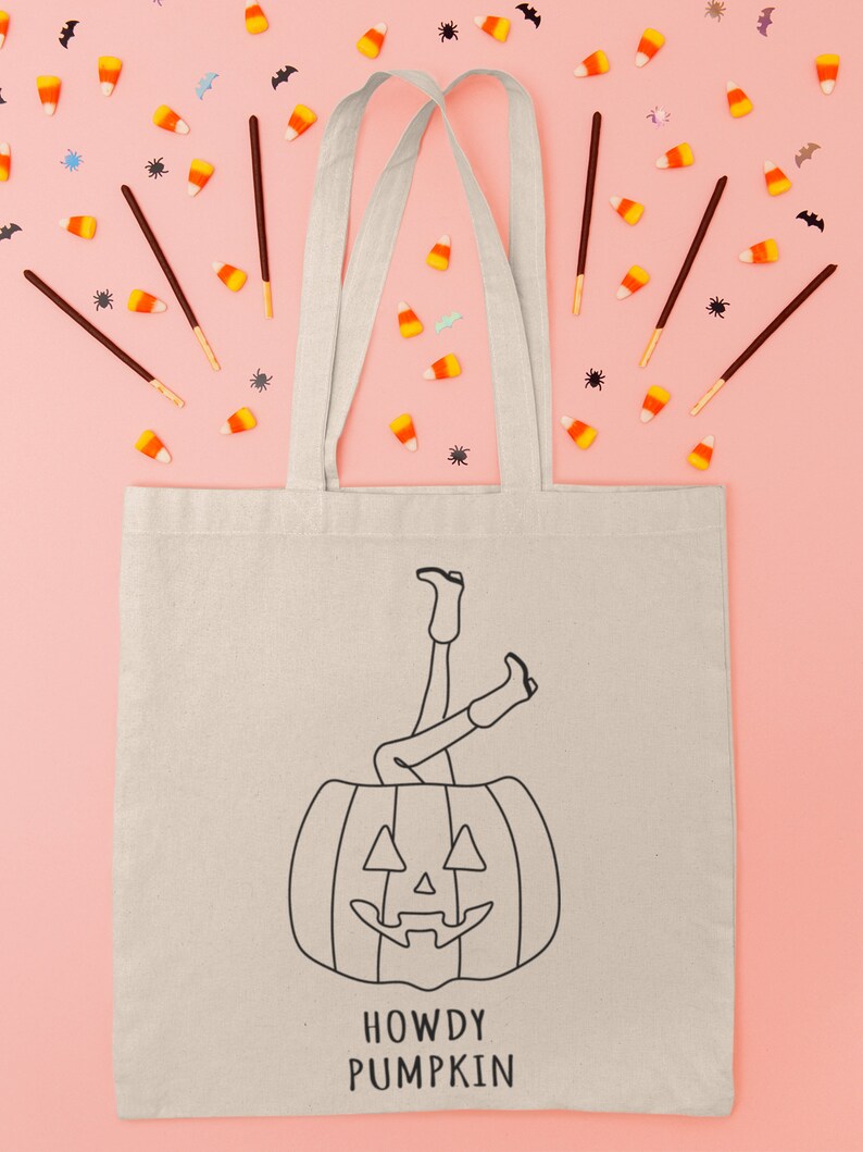 May include: A white canvas tote bag with black text that says "Howdy Pumpkin" and a black line drawing of a person's legs sticking out of a jack-o'-lantern. The bag is surrounded by candy corn, bats, and spiders on a pink background.