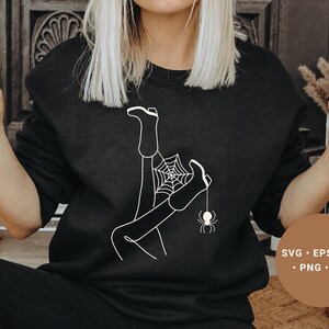 May include: A black sweatshirt with a white graphic design of a person's legs tangled in a spider web with a spider hanging from the web. The text "SVG - EPS - DXF - PNG - JPG" is printed on a brown tag.