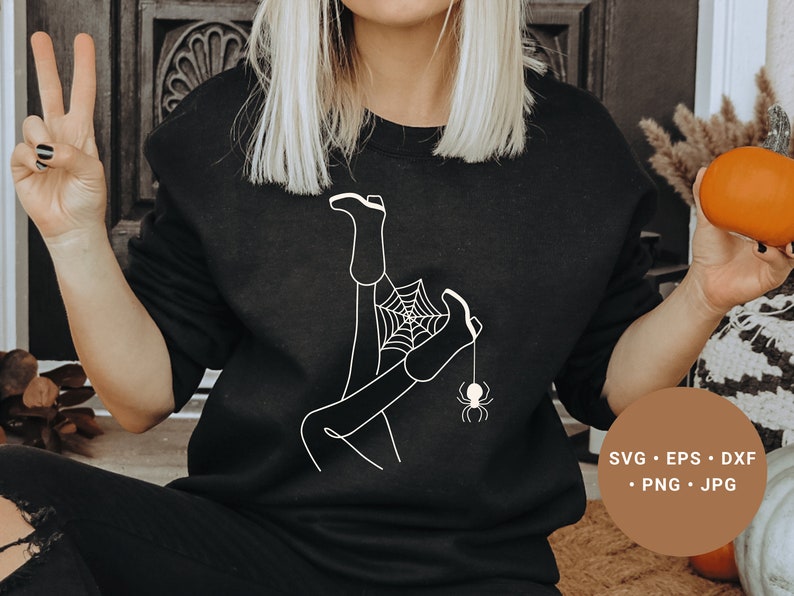 May include: A black sweatshirt with a white graphic design of a person's legs tangled in a spider web with a spider hanging from the web. The text "SVG - EPS - DXF - PNG - JPG" is printed on a brown tag.