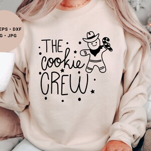 May include: A beige sweatshirt with the text "THE cookie CREW" in black lettering. A gingerbread man with a cowboy hat and candy cane is holding a star.