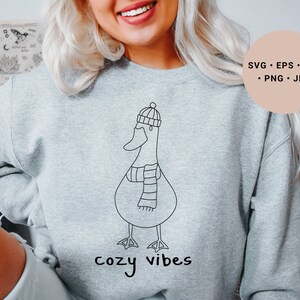 K&ouml;nnte beinhalten: Ein graues Sweatshirt mit einer schwarzen Strichzeichnung eines Entchens, das einen Hut und einen Schal tr&auml;gt. Der Text "cozy vibes" ist unter dem Entchen gedruckt.