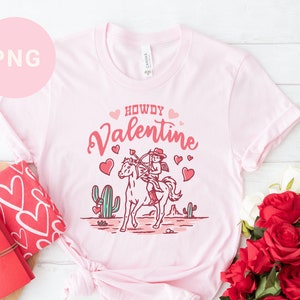 May include: Pink t-shirt with a cowboy riding a horse and the text "Howdy Valentine" in pink lettering with hearts.