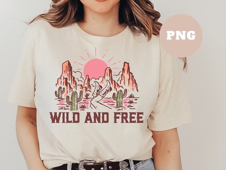 Puede incluir: Una camiseta beige con un gr&aacute;fico de un paisaje des&eacute;rtico con monta&ntilde;as, un camino y cactus. El sol se est&aacute; poniendo en el fondo. El texto "WILD AND FREE" est&aacute; impreso debajo del gr&aacute;fico.