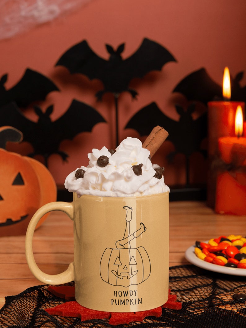 May include: A beige ceramic mug with a black and white illustration of a pumpkin with legs sticking out of the top. The mug has the text "Howdy Pumpkin" on the bottom. The mug is filled with whipped cream and chocolate chips and topped with a cinnamon stick. The mug is sitting on a black and white lace doily.
