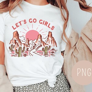 May include: White t-shirt with a graphic of a desert landscape with a pink sun, cacti, and the text "Let's Go Girls".