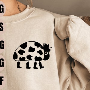 Könnte beinhalten: Ein schwarz-weißes Kuhflecken-Marienkäfer, das Cowboystiefel trägt. Der Marienkäfer trägt einen Cowboyhut mit einem Stern oben drauf. Der Marienkäfer befindet sich auf einem hellbraunen Sweatshirt.