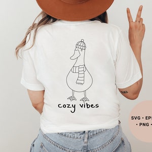 K&ouml;nnte beinhalten: Wei&szlig;es T-Shirt mit einer schwarzen Strichzeichnung eines Entchens, das einen Hut und einen Schal tr&auml;gt. Der Text "cozy vibes" ist unter dem Entchen gedruckt.