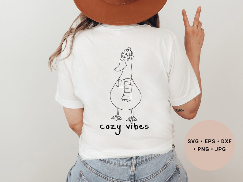 K&ouml;nnte beinhalten: Wei&szlig;es T-Shirt mit einer schwarzen Strichzeichnung eines Entchens, das einen Hut und einen Schal tr&auml;gt. Der Text "cozy vibes" ist unter dem Entchen gedruckt.