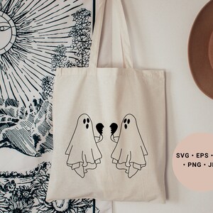May include: A white tote bag featuring two black ghosts holding broken hearts. The bag is hanging on a white wall with a black and white tapestry and a brown hat in the background. The text "SVG - EPS - DXF - PNG - JPG" is printed on a pink circle in the bottom right corner of the image.
