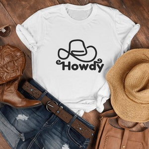 Może przedstawiać: Biała koszulka z czarnym rysunkiem kowbojskiego kapelusza i nadrukiem słowa "Howdy". Koszulka leży na drewnianej powierzchni z parą brązowych kowbojskich but&oacute;w, dżinsową sp&oacute;dnicą, brązowym paskiem, słomkowym kapeluszem i brązową sk&oacute;rzaną torbą.