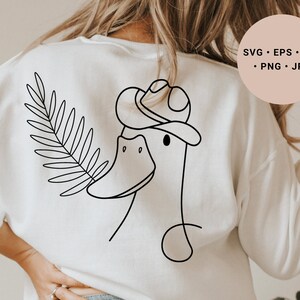 Könnte beinhalten: Ein weißes Sweatshirt mit einer schwarzen Linienzeichnung eines Entchens, das einen Cowboyhut und ein Palmblatt trägt. Der Text "SVG - EPS - DXF - PNG - JPG" befindet sich auf einem rosa Rechteck in der oberen rechten Ecke des Bildes.