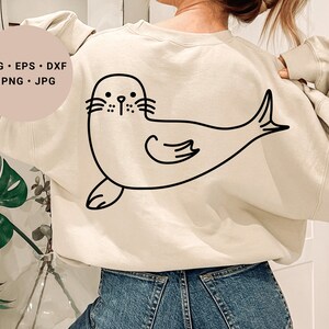 May include: A cream-colored sweatshirt with a black line drawing of a seal on the back. The seal is facing the left side of the image and has a simple, cartoon-like design.